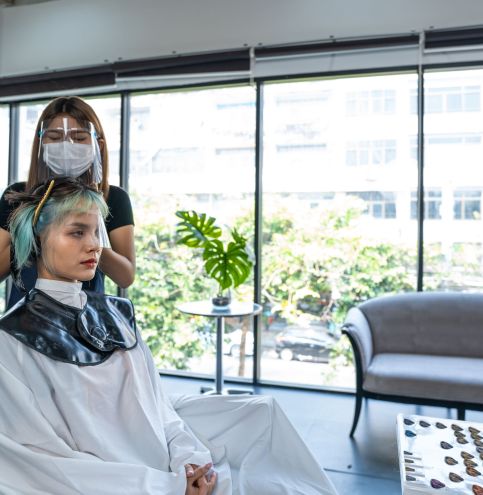 Professional woman hairstylist dyeing young woman customer's hair or change color in beauty salon. Hairdresser wearing face shield to prevent from coronavirus infection. Beauty salon business concept.